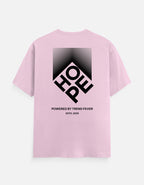 A light pink short-sleeve t-shirt shown from the back, featuring a large black graphic print of the word 'HOPE' inside a diamond shape with a gradient background. Below the graphic, the text reads 'POWERED BY TREND FEVER' and 'ESTD.