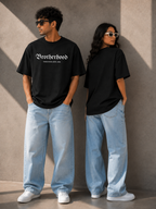 Two models wearing matching black t-shirts with 'Brotherhood' printed in white Gothic font, paired with light-wash jeans and white sneakers, standing against a textured concrete wall.