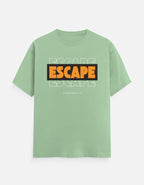 A sage green short-sleeve t-shirt with a graphic on the chest reading 'ESCAPE' in orange and black, layered over faint white 'ESCAPE' text and 'FROM REALITY' below, shown on a light gray background.