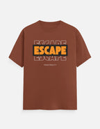 A brown short-sleeve t-shirt with a graphic on the chest reading 'ESCAPE' in orange and black, with 'FROM REALITY' printed below, displayed on a light gray background.