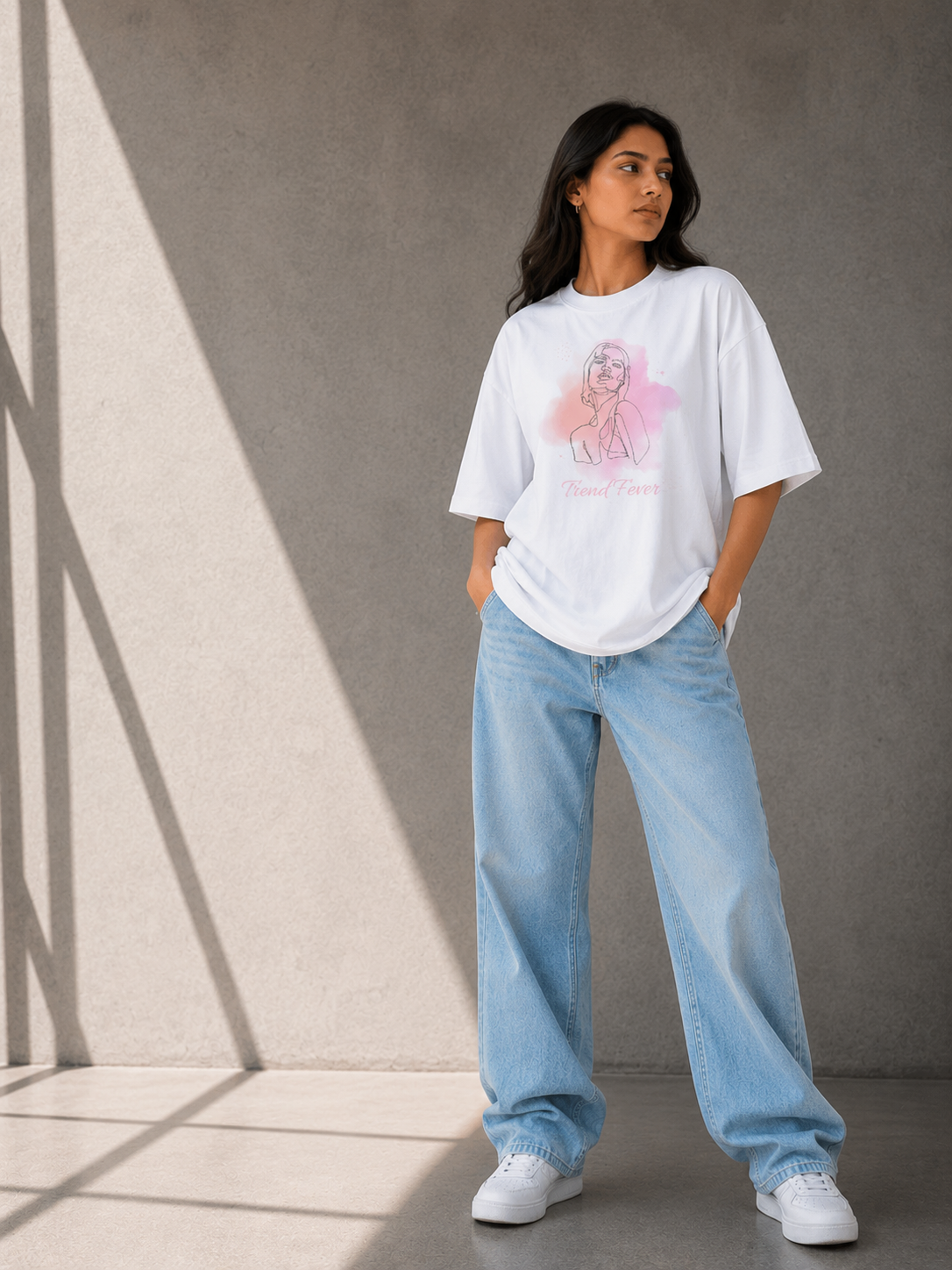 A woman stands against a textured gray concrete wall wearing a TREND FEVER white oversized graphic T-shirt with a pink and purple illustration, light blue wide-leg jeans, and white sneakers.