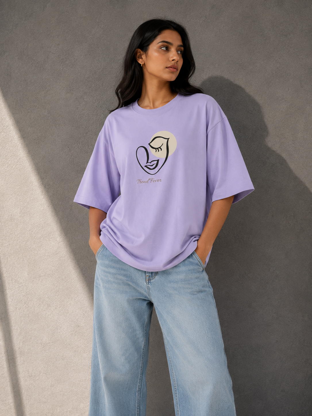 A woman modeling an TREND FEVER oversized light purple T-shirt with a minimalist line art graphic of a face and heart on the front, paired with light blue wide-leg jeans, standing against a textured gray wall.