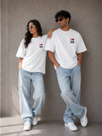 A man and a woman wearing Trend Fever matching white graphic t-shirts with a small red heart and black text reading 'I [heart] ME' on the left chest, paired with light-wash jeans and white sneakers, posing against a textured gray wall.