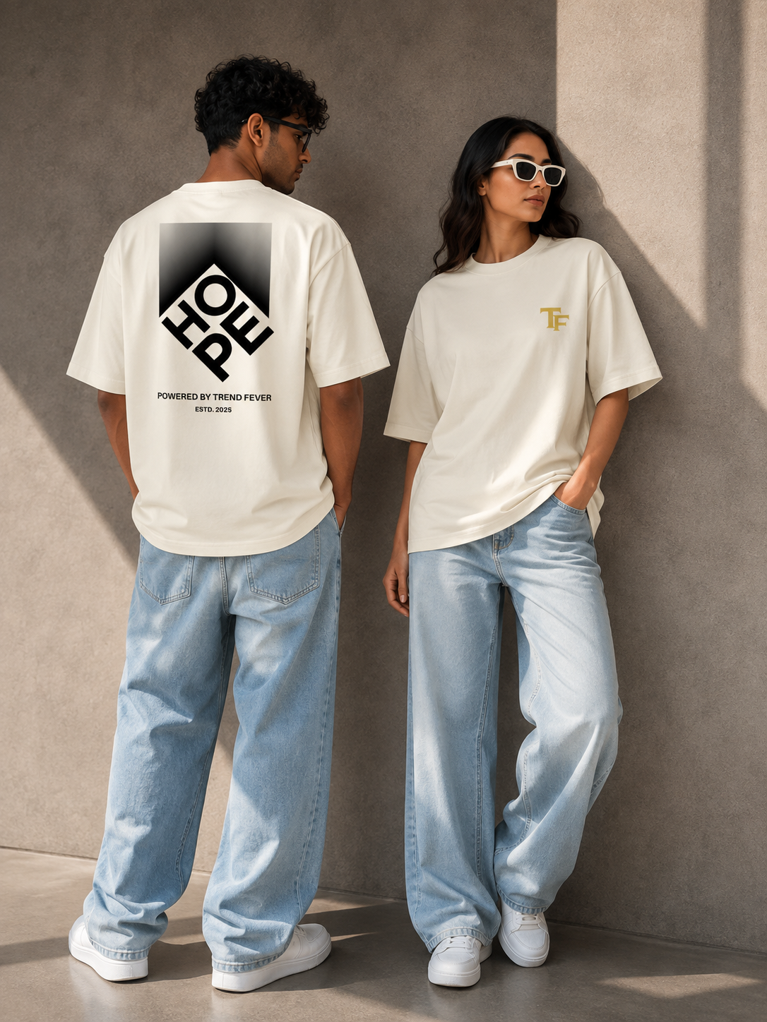 Two models wearing off-white TREND FEVER oversized T-shirts and light-wash jeans standing against a textured concrete wall. The male model shows the back of the shirt with a large black geometric 'HOPE' graphic, and the female model shows the front with a small gold 'TF' logo on the chest.