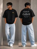 A man modeling a black oversized T-shirt shown from both front and back against a concrete wall. The front has a small gold chest logo, and the back has a large white graphic reading 'Galford' with smaller text below.