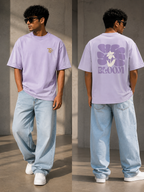 A man modeling a light purple oversized t-shirt, shown from both the front and back. The front features a small gold 'TF' logo on the left chest, while the back displays a large purple flower graphic with a white hand in the center and the word 'BLOOM' in purple.