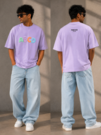 A man modeling a light purple t-shirt from both front and back views, paired with light blue jeans and white sneakers against a textured concrete wall.