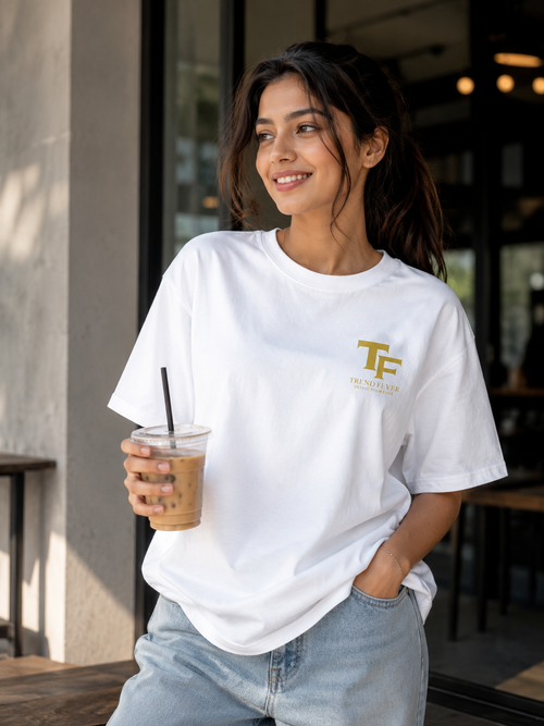 A woman wearing a white short-sleeve t-shirt with a gold 'TREND FEVER' logo and 'THE FUTURE OF YOU' text on the left chest, holding an iced coffee in front of a cafe setting.
