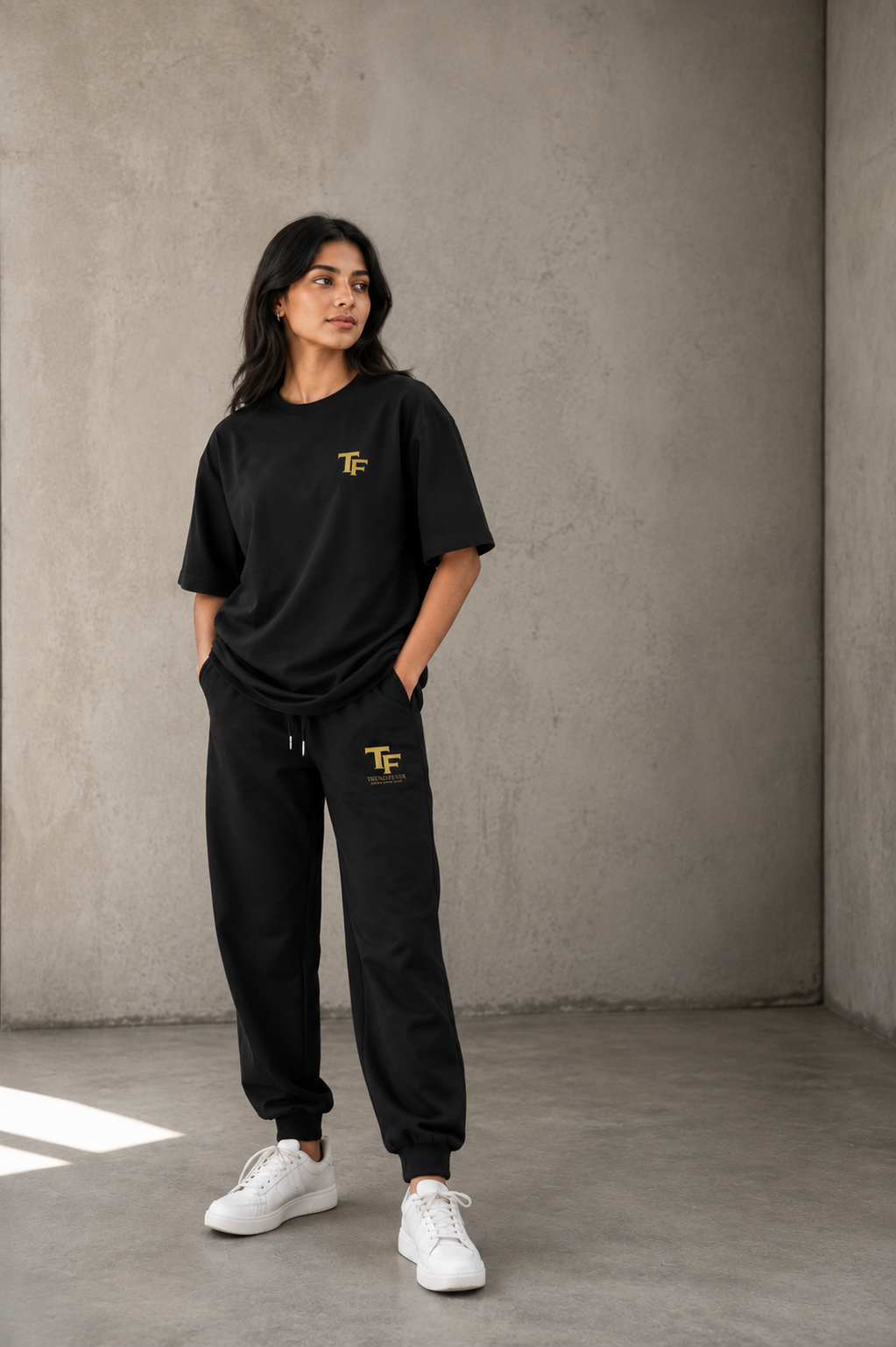 A woman modeling a black oversized t-shirt and matching black jogger sweatpants with a small yellow 'TF' logo on the chest and thigh, paired with white sneakers, standing against a textured concrete wall.