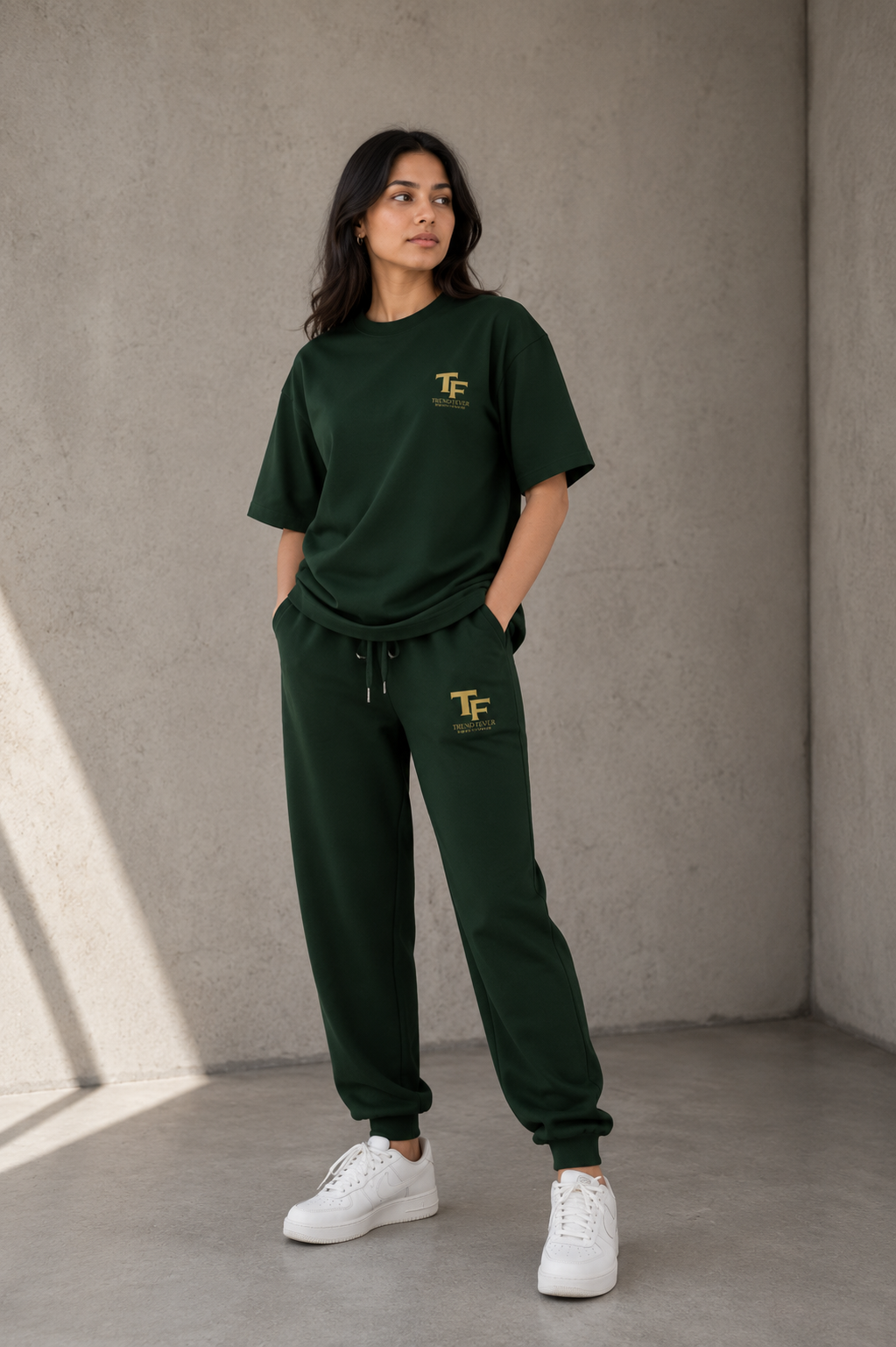A woman modeling a dark green matching lounge set consisting of a short-sleeve t-shirt and jogger pants, both featuring a gold 'TF' monogram logo on the chest and upper thigh. She is wearing white sneakers and standing in a minimalist concrete room corner.
