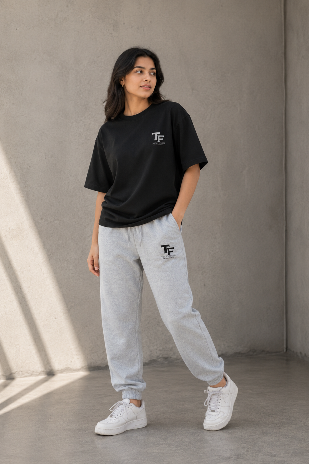 A woman modeling a black oversized t-shirt and light gray sweatpants with a matching 'TF' logo, paired with white sneakers, standing against a textured concrete wall.