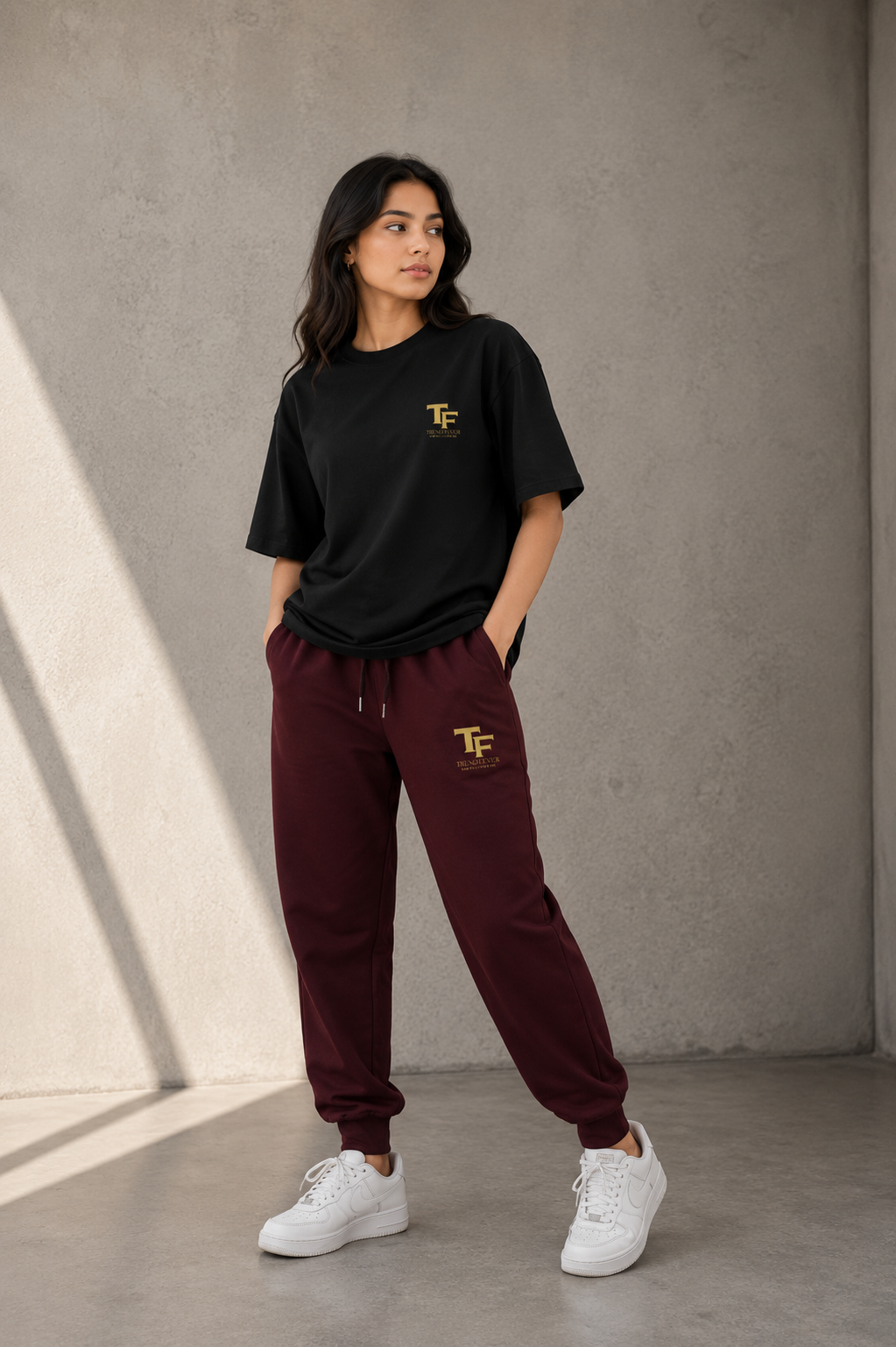 A woman modeling a black crewneck t-shirt and burgundy jogger sweatpants, both featuring a small gold 'TF' logo on the chest and thigh, paired with white sneakers against a textured concrete wall.