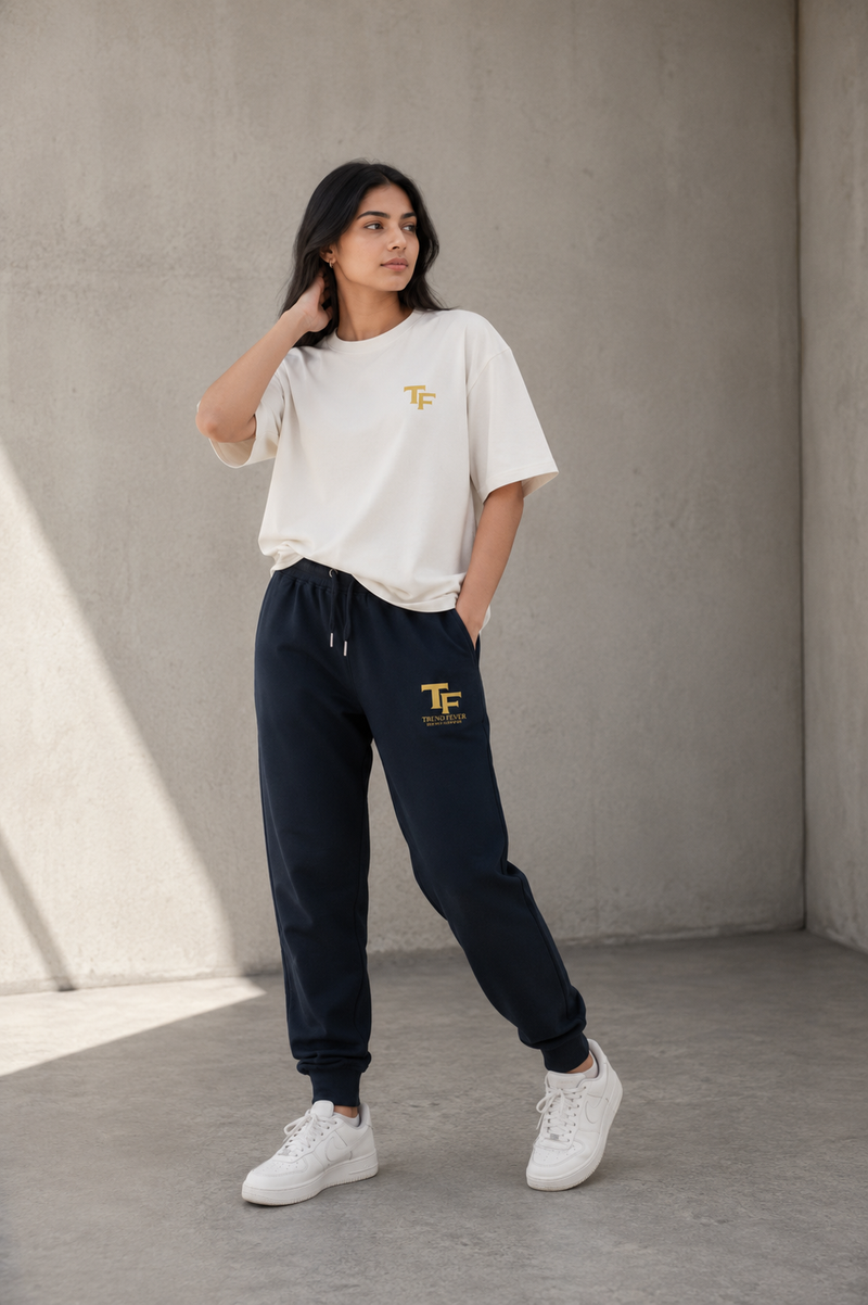 A woman modeling a white short-sleeve t-shirt and navy jogger sweatpants with a yellow 'TF' logo, standing against a textured concrete wall.