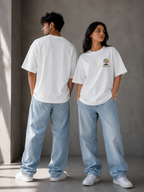 Two models wearing Trend Fever white oversized t-shirts and light blue jeans standing in a minimalist concrete room. The model on the left shows the back of the shirt, while the model on the right shows the front, which features a small colorful circular graphic on the left chest.