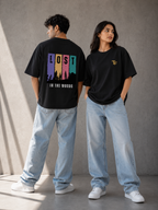 Two models wearing black graphic t-shirts and light-wash jeans standing against a textured gray wall. The model on the left shows the back of the t-shirt with a colorful 'LOST IN THE WOODS' graphic, while the model on the right shows the front with a small gold 'TF' logo on the chest.